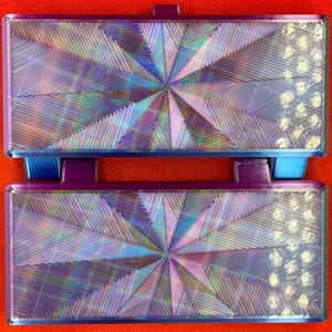 May include: Two rectangular, hinged components featuring a vibrant, iridescent finish with a central starburst pattern of fine, radiating lines. The edges are framed in a deep purple, and the right side displays a textured, dotted pattern.