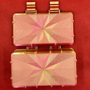 May include: Two stacked pink and gold rectangular boxes with a geometric starburst design. The boxes have a textured surface and are made of a hard plastic material.