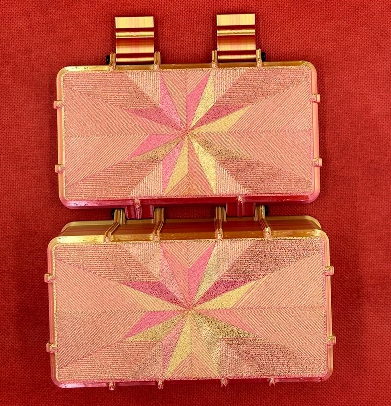 May include: Two stacked pink and gold rectangular boxes with a geometric starburst design. The boxes have a textured surface and are made of a hard plastic material.