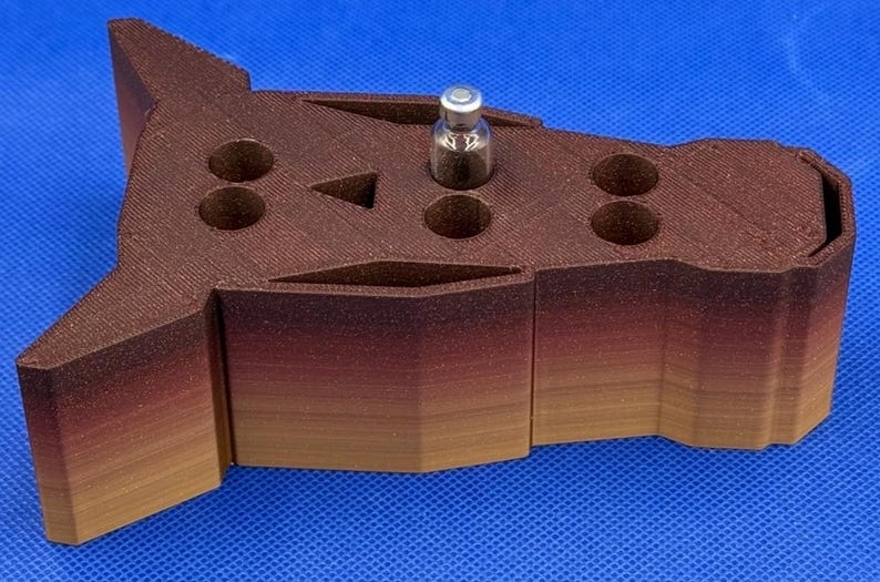 May include: A 3D printed, brown and tan animal head shaped organizer with multiple holes for holding small items. A small glass vial is sitting in one of the holes.