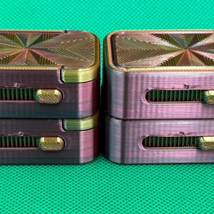 May include: Four rectangular, metallic-finish containers stacked in two pairs. Each container features a top surface with a geometric, starburst-like pattern and a side-mounted sliding mechanism with a gold-coloured textured switch.