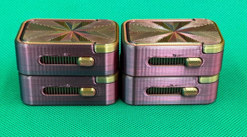 May include: Four rectangular, metallic-finish containers stacked in two pairs. Each container features a top surface with a geometric, starburst-like pattern and a side-mounted sliding mechanism with a gold-coloured textured switch.