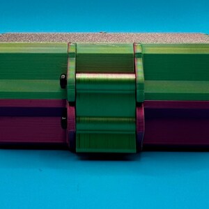 Puede incluir: Una caja de almacenamiento rectangular con un dise&ntilde;o de dos tonos, que presenta una secci&oacute;n superior de color verde met&aacute;lico y una secci&oacute;n inferior de color morado oscuro. El contenedor incluye un mecanismo de cierre central y dos peque&ntilde;os cierres negros.