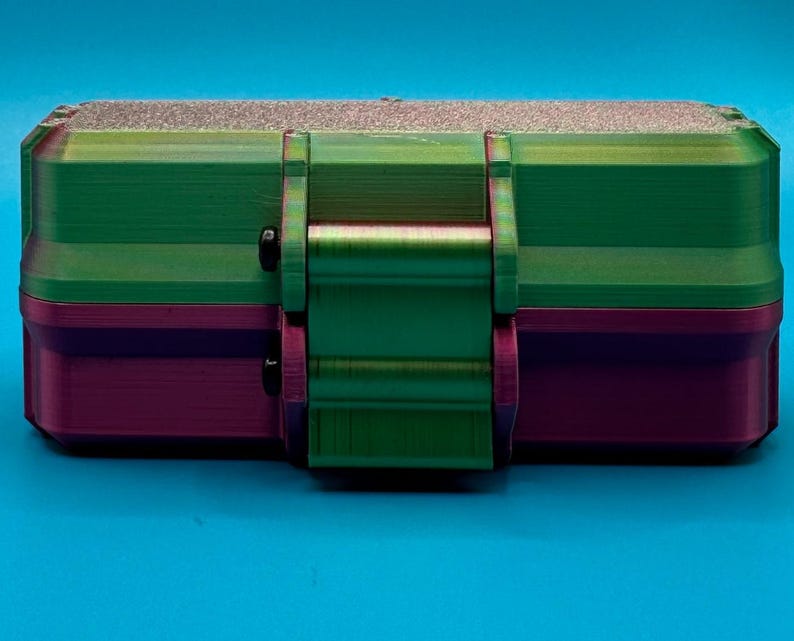 Puede incluir: Una caja de almacenamiento rectangular con un dise&ntilde;o de dos tonos, que presenta una secci&oacute;n superior de color verde met&aacute;lico y una secci&oacute;n inferior de color morado oscuro. El contenedor incluye un mecanismo de cierre central y dos peque&ntilde;os cierres negros.