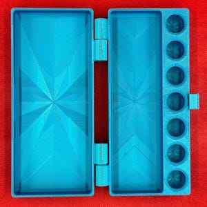 May include: A bright blue, rectangular, hinged plastic case featuring a large open compartment on the left and a matching compartment on the right, accompanied by a vertical column of seven circular wells for holding small items.
