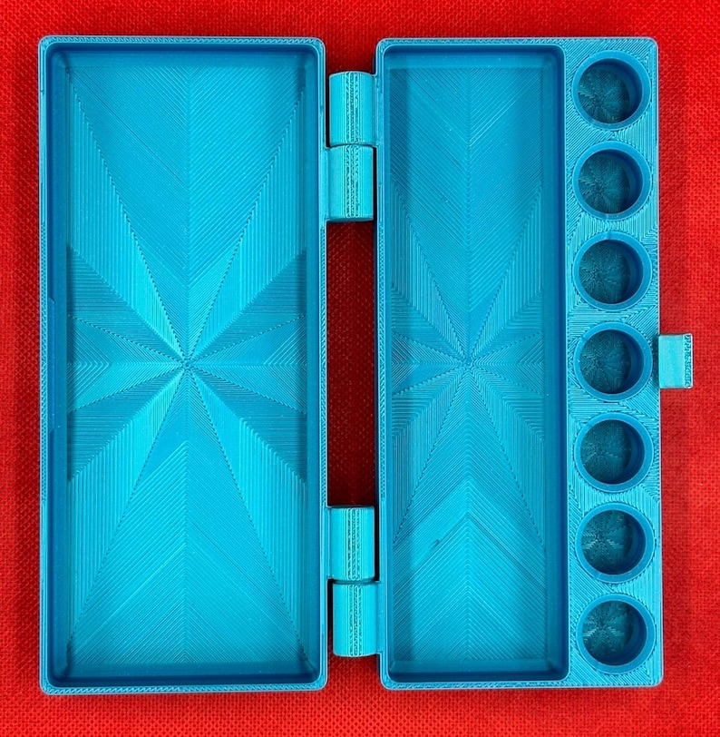 May include: A bright blue, rectangular, hinged plastic case featuring a large open compartment on the left and a matching compartment on the right, accompanied by a vertical column of seven circular wells for holding small items.