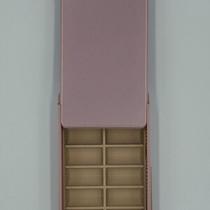 May include: A rectangular, open, pink jewelry organizer with multiple small compartments. The lid is open, revealing the interior compartments. The organizer is made of a textured material and is designed to store small items like jewelry.