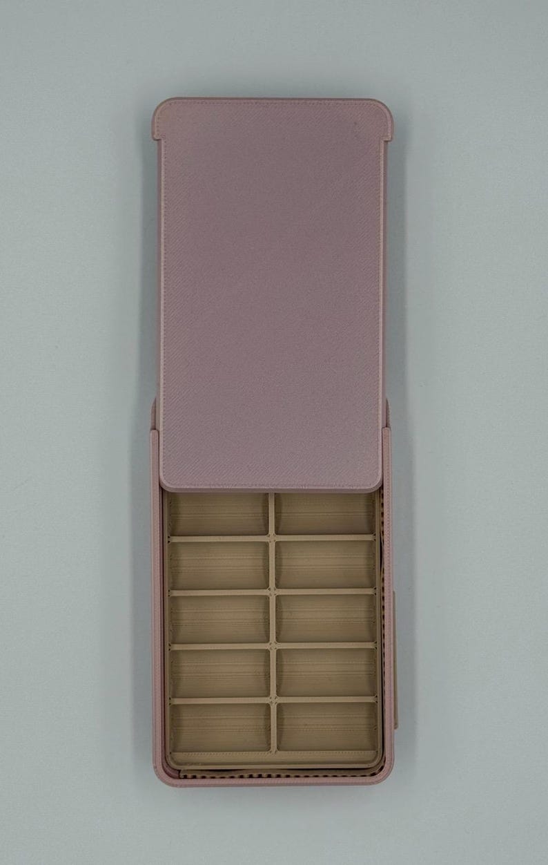May include: A rectangular, open, pink jewelry organizer with multiple small compartments. The lid is open, revealing the interior compartments. The organizer is made of a textured material and is designed to store small items like jewelry.