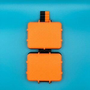 May include: A rectangular orange storage case with a black hinge and latch mechanism. The case features a textured surface and a small black and orange tab extending from the top edge, designed for secure closure and organisation of small items.