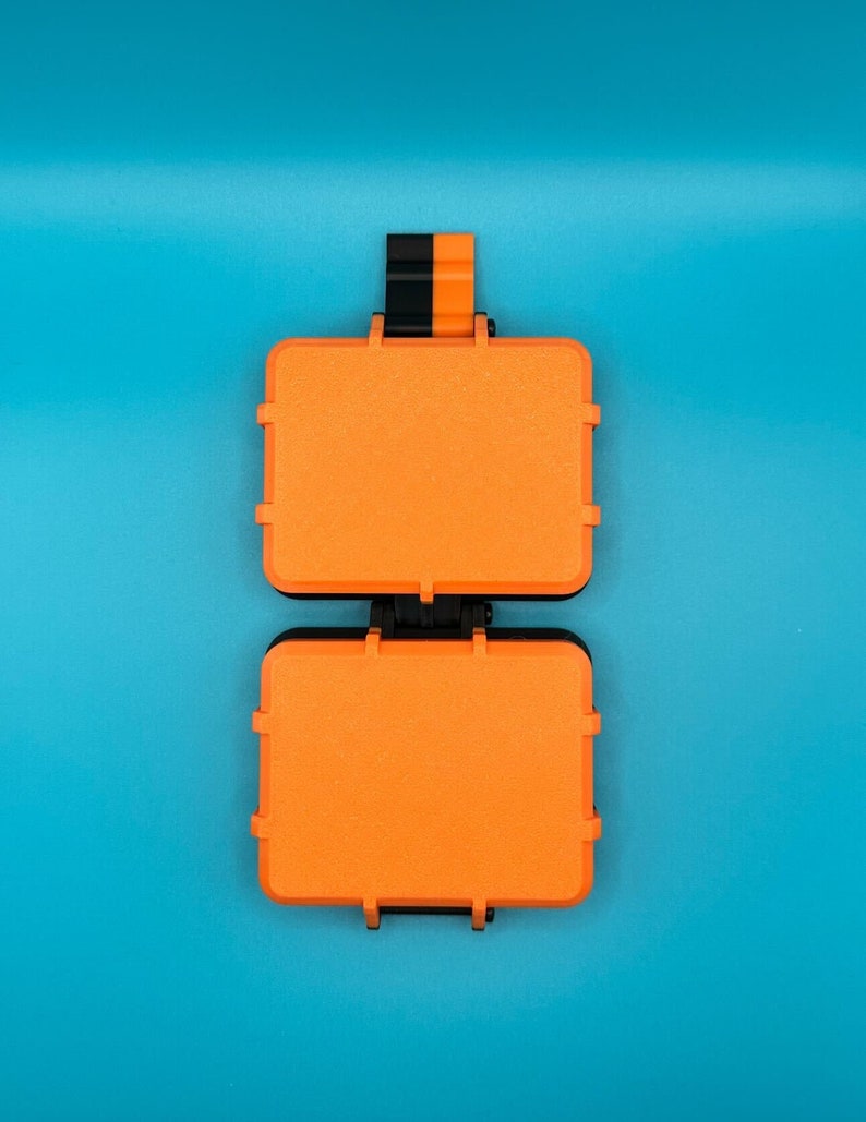 May include: A rectangular orange storage case with a black hinge and latch mechanism. The case features a textured surface and a small black and orange tab extending from the top edge, designed for secure closure and organisation of small items.