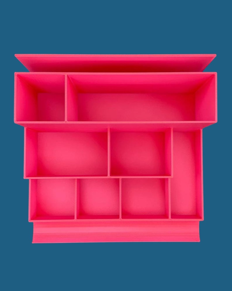 May include: A bright hot pink desk organizer featuring a tiered design with multiple rectangular compartments of varying sizes. The structure includes one long top section, two middle sections, and four smaller bottom sections for storage.