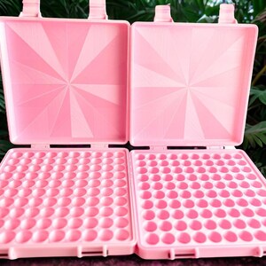 May include: Two open, square, light pink plastic waffle makers with a grid of small circular indentations on the bottom plates and a sunburst pattern embossed on the interior of the top lids, featuring hinged closures at the top edges.
