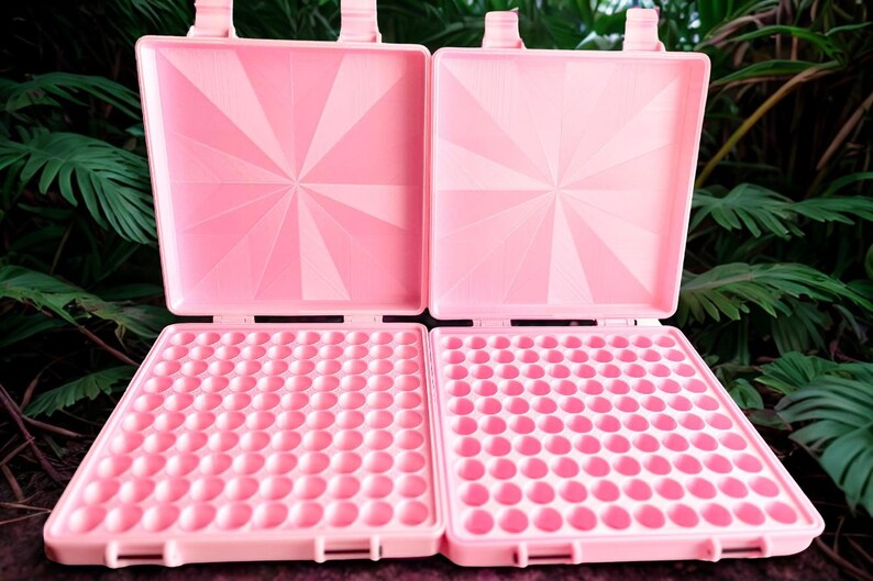 May include: Two open, square, light pink plastic waffle makers with a grid of small circular indentations on the bottom plates and a sunburst pattern embossed on the interior of the top lids, featuring hinged closures at the top edges.