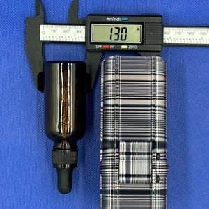 May include: A dark amber glass dropper bottle with a black cap and a black digital caliper measuring 1.30 inches in width. The bottle is positioned next to a rectangular object featuring a black and white horizontal striped pattern.