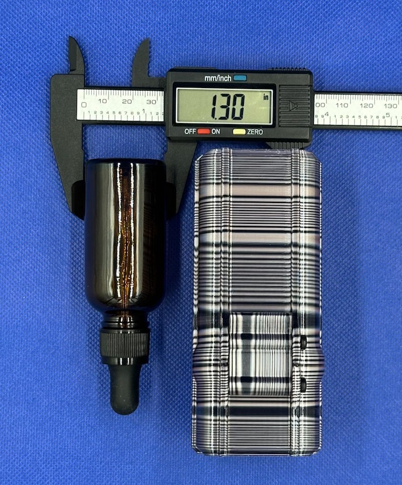 May include: A dark amber glass dropper bottle with a black cap and a black digital caliper measuring 1.30 inches in width. The bottle is positioned next to a rectangular object featuring a black and white horizontal striped pattern.