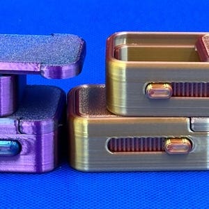 May include: Two sets of rectangular fidget sliders, one purple and one gold, each featuring a textured sliding top plate and a side-mounted mechanical switch mechanism. The objects are stacked in pairs against a solid blue background.