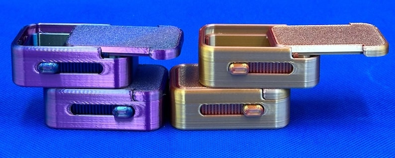 May include: Two sets of rectangular fidget sliders, one purple and one gold, each featuring a textured sliding top plate and a side-mounted mechanical switch mechanism. The objects are stacked in pairs against a solid blue background.
