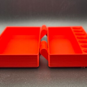 May include: A bright red rectangular plastic storage case with a hinged lid. The interior features a large open compartment on the left side and a smaller rectangular compartment with six circular indentations on the right side.