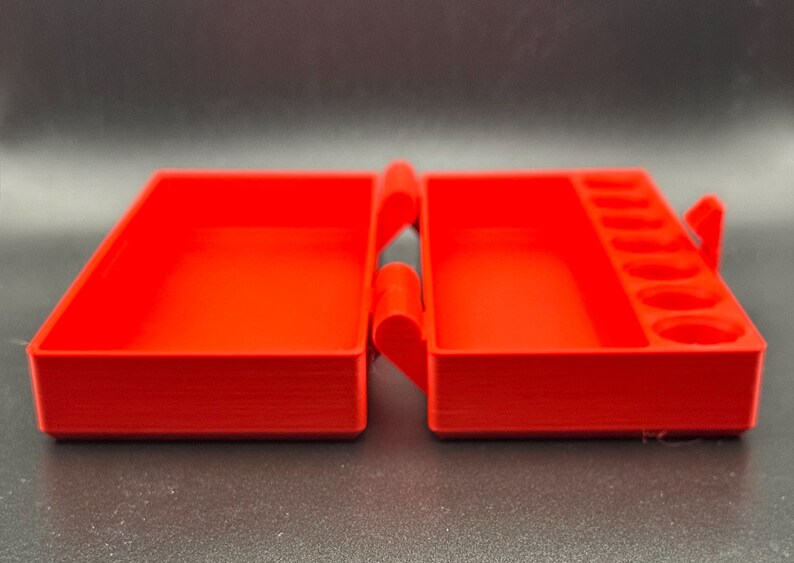 May include: A bright red rectangular plastic storage case with a hinged lid. The interior features a large open compartment on the left side and a smaller rectangular compartment with six circular indentations on the right side.