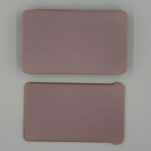 May include: Two rectangular, dusty rose-colored tablet cases. The top case is closed, while the bottom case is open, revealing a slightly different shape. Both cases have rounded corners and a textured surface.