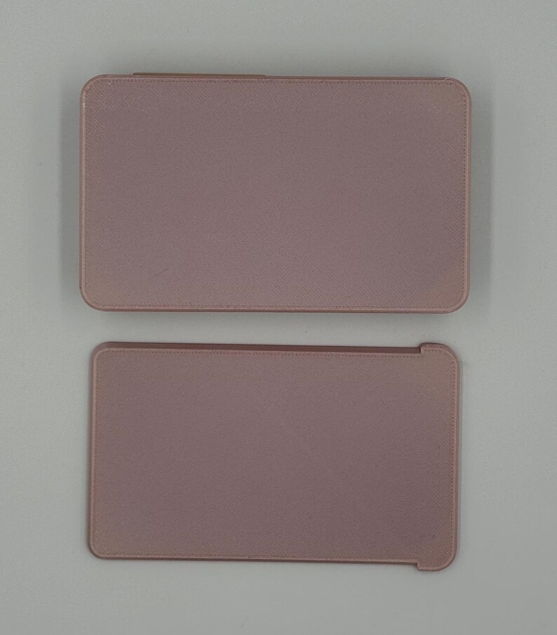 May include: Two rectangular, dusty rose-colored tablet cases. The top case is closed, while the bottom case is open, revealing a slightly different shape. Both cases have rounded corners and a textured surface.