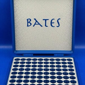 K&ouml;nnte beinhalten: Eine blaue Plastikbox mit grauem Innenraum und einem Raster aus 64 runden F&auml;chern. Die Box hat einen silbernen Verschluss und das Wort "BATES" ist auf dem Deckel aufgedruckt.