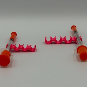 May include: Two orange and clear syringes with pink holders. The syringes have orange caps and are positioned on a white surface. The pink holders have a wavy design with multiple slots.