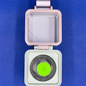 May include: A square, hinged container with a light pink lid and a white base. The interior of the base features a circular cutout containing a bright lime green circular insert. The container has a visible hinge mechanism at the top.