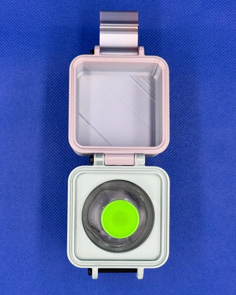May include: A square, hinged container with a light pink lid and a white base. The interior of the base features a circular cutout containing a bright lime green circular insert. The container has a visible hinge mechanism at the top.