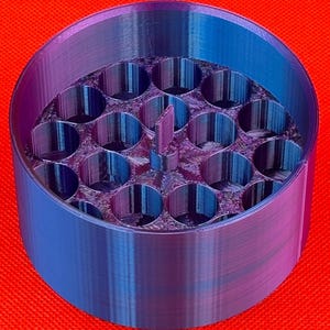 May include: A cylindrical container with a gradient blue and purple finish, featuring a circular interior grid with nineteen vertical tube-like compartments and a small central rectangular tab extending upward from the base.