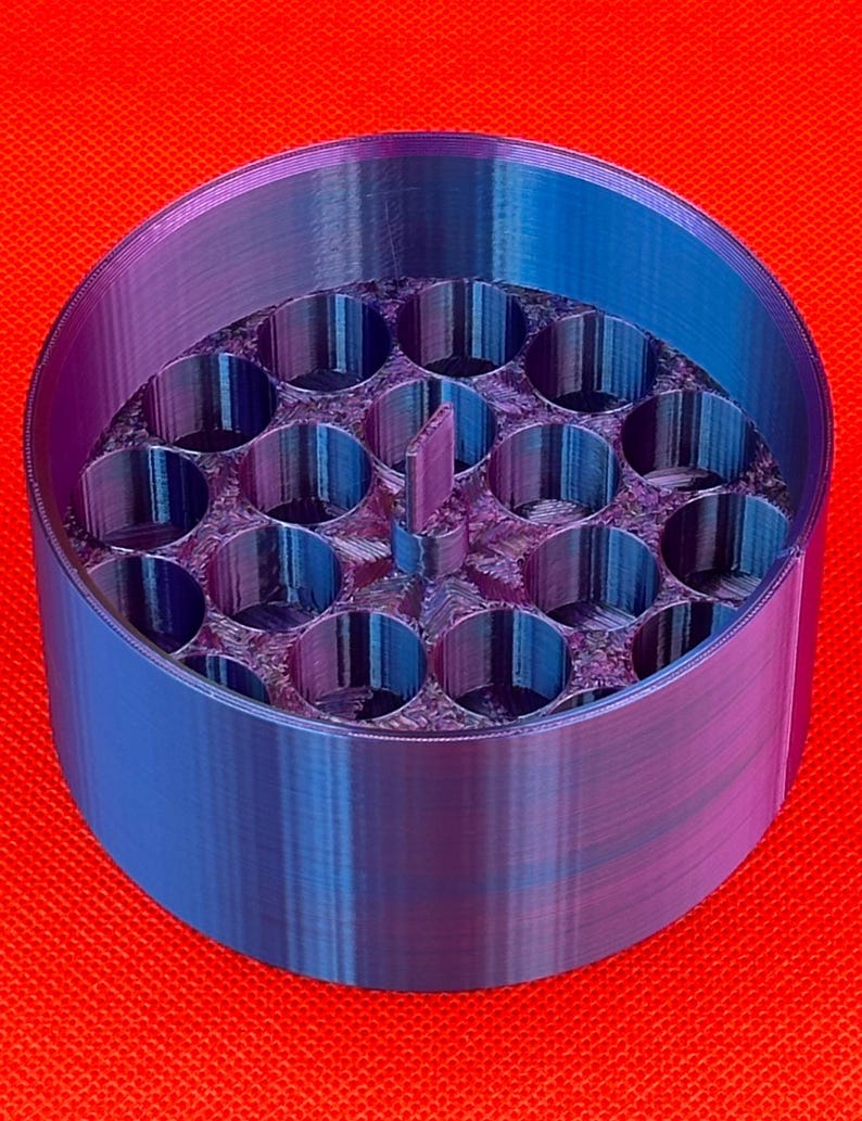 May include: A cylindrical container with a gradient blue and purple finish, featuring a circular interior grid with nineteen vertical tube-like compartments and a small central rectangular tab extending upward from the base.