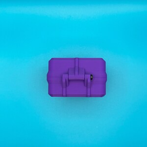 May include: A rectangular purple storage case featuring a central latch mechanism with a small black pin detail. The case has a matte finish and a structured, boxy design with reinforced edges, presented from a top-down perspective.