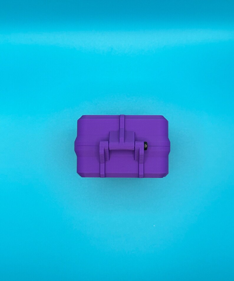 May include: A rectangular purple storage case featuring a central latch mechanism with a small black pin detail. The case has a matte finish and a structured, boxy design with reinforced edges, presented from a top-down perspective.
