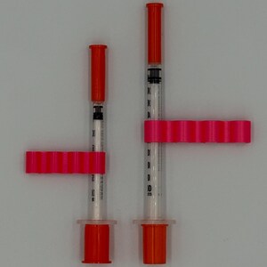 May include: Two medical syringes with orange caps and pink holders. The syringes are clear with black markings for measurement. The pink holders are rectangular with multiple rounded sections. The syringes are on a white background.