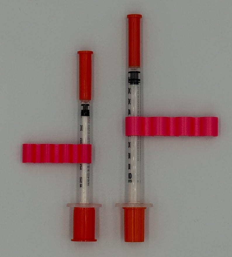 May include: Two medical syringes with orange caps and pink holders. The syringes are clear with black markings for measurement. The pink holders are rectangular with multiple rounded sections. The syringes are on a white background.