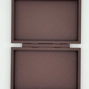 May include: Open, rectangular brown box with a hinged lid. The box is made of a solid material and has a textured surface. The lid is open, revealing the interior of the box. The box is sitting on a white surface.