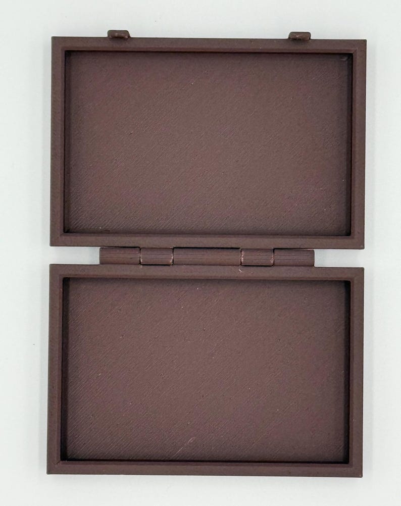May include: Open, rectangular brown box with a hinged lid. The box is made of a solid material and has a textured surface. The lid is open, revealing the interior of the box. The box is sitting on a white surface.