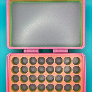 May include: A rectangular storage case with a pink outer frame and a rainbow-colored interior rim. The top lid features a flat gray panel, while the bottom section contains a grid of thirty-five circular cutouts with a peach-colored base.