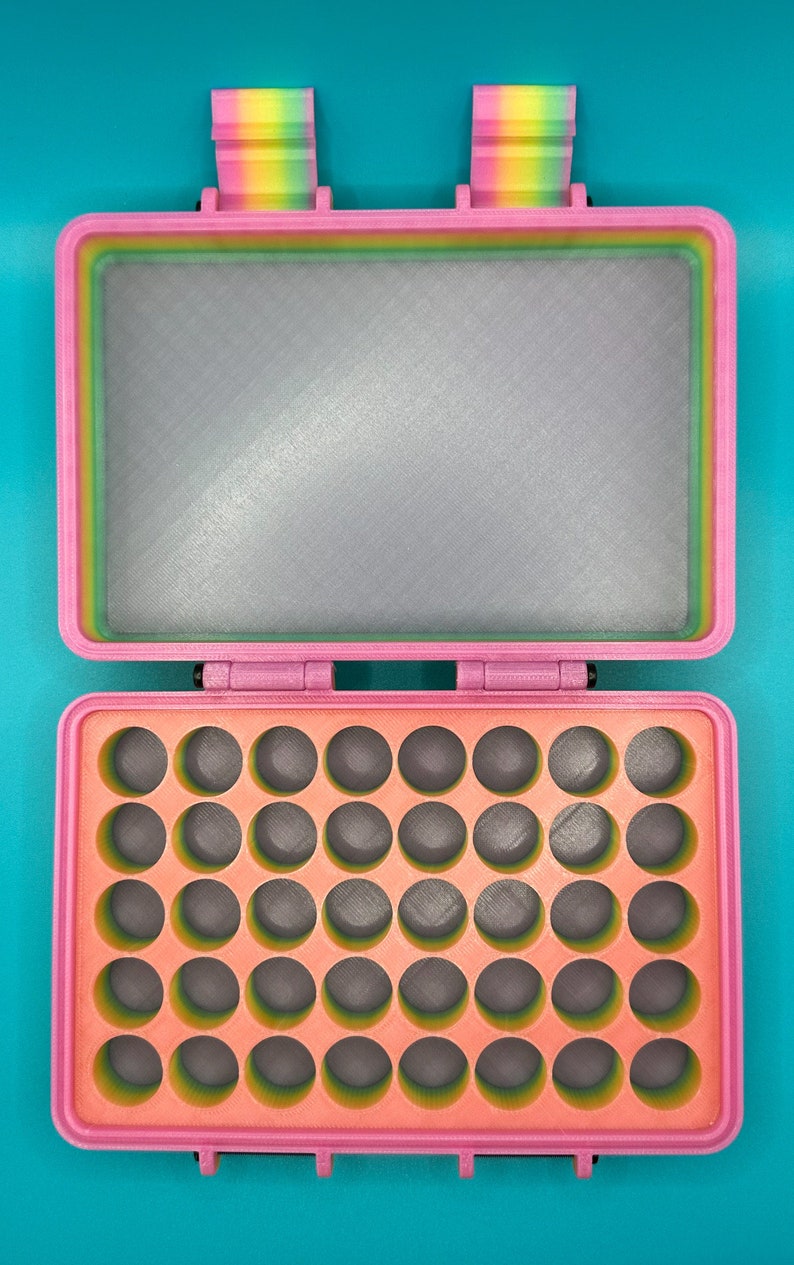 May include: A rectangular storage case with a pink outer frame and a rainbow-colored interior rim. The top lid features a flat gray panel, while the bottom section contains a grid of thirty-five circular cutouts with a peach-colored base.