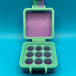 May include: A mint green square storage case with a hinged lid and a gradient pink and green latch. The interior features a grid of nine circular compartments with a purple base. The case has a black metal hinge pin at the front closure.