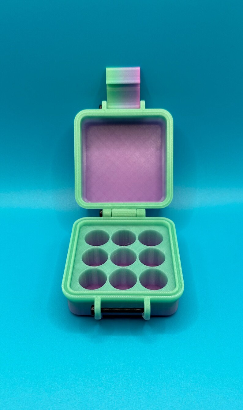 May include: A mint green square storage case with a hinged lid and a gradient pink and green latch. The interior features a grid of nine circular compartments with a purple base. The case has a black metal hinge pin at the front closure.