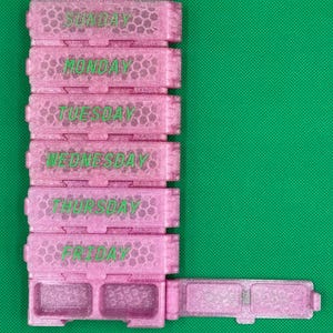 May include: A pink glittery weekly pill organizer with a honeycomb texture. The stack features individual compartments labeled Sunday through Friday in green text. The bottom section has two open compartments with a hinged lid extended to the right side.