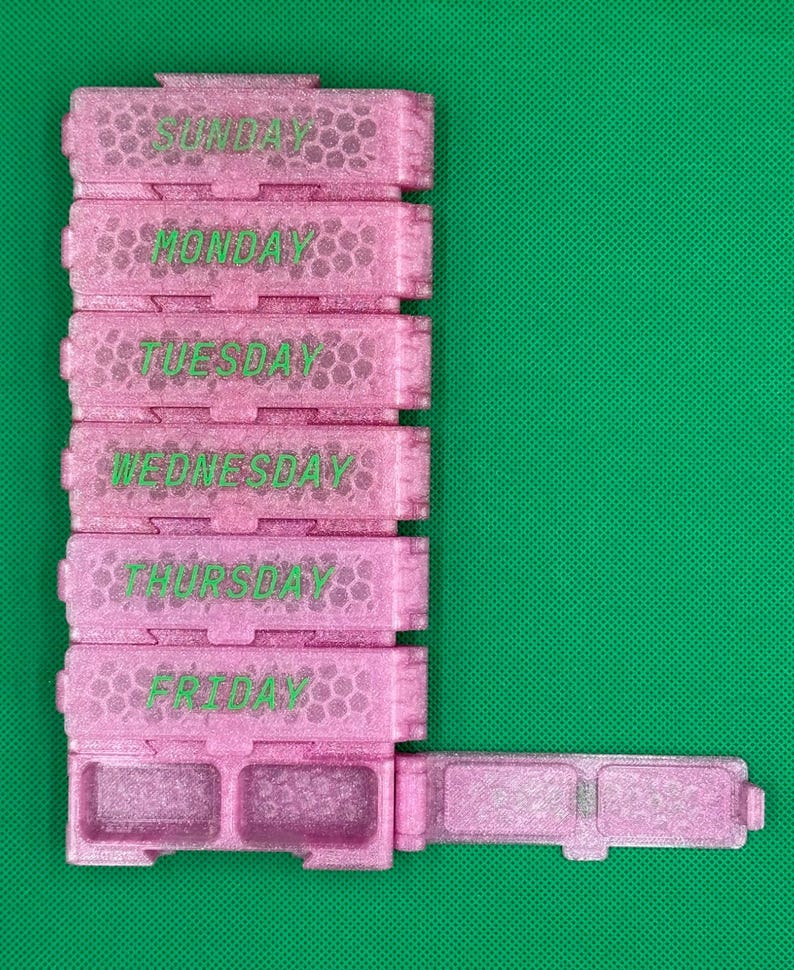 May include: A pink glittery weekly pill organizer with a honeycomb texture. The stack features individual compartments labeled Sunday through Friday in green text. The bottom section has two open compartments with a hinged lid extended to the right side.