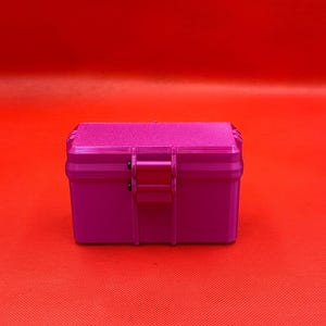 May include: A rectangular, magenta-colored storage box with a textured lid and a central latch mechanism. The box features a smooth, solid-colored body and a slightly rougher, matte-finish top surface, secured by a hinged closure.