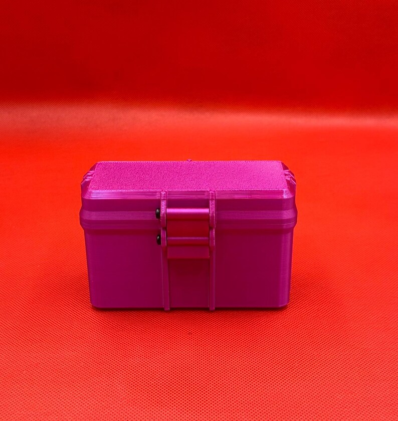 May include: A rectangular, magenta-colored storage box with a textured lid and a central latch mechanism. The box features a smooth, solid-colored body and a slightly rougher, matte-finish top surface, secured by a hinged closure.