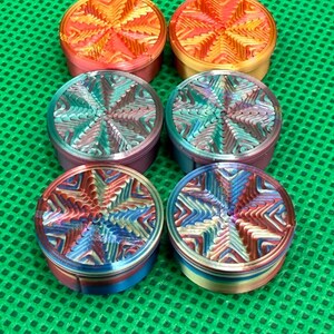 May include: Six circular metal herb grinders arranged in two columns of three. Each grinder features a distinct, multi-colored gradient finish with a textured, star-shaped geometric pattern on the lid, displaying vibrant hues of red, orange, blue, and purple.