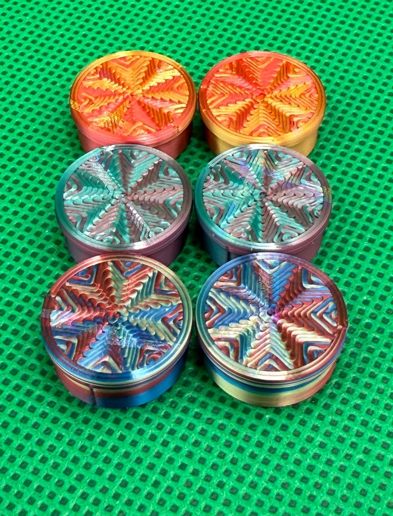 May include: Six circular metal herb grinders arranged in two columns of three. Each grinder features a distinct, multi-colored gradient finish with a textured, star-shaped geometric pattern on the lid, displaying vibrant hues of red, orange, blue, and purple.