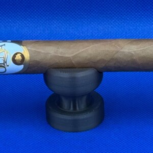May include: A brown cigar with a white and gold label that reads "OLIVA" is resting on a black cigar holder.