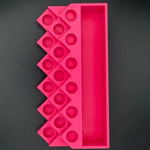 May include: A bright pink organizer tray featuring a long rectangular compartment on one side and a series of sixteen circular indentations arranged in a zigzag pattern on the other side, constructed with a matte finish.