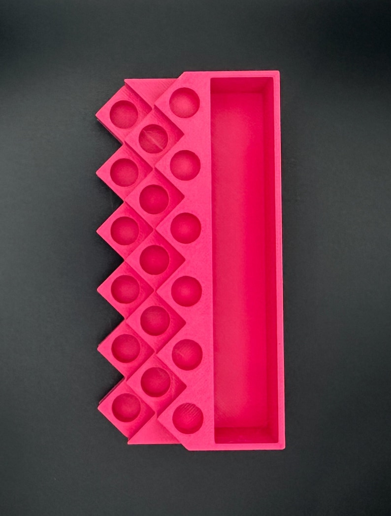 May include: A bright pink organizer tray featuring a long rectangular compartment on one side and a series of sixteen circular indentations arranged in a zigzag pattern on the other side, constructed with a matte finish.